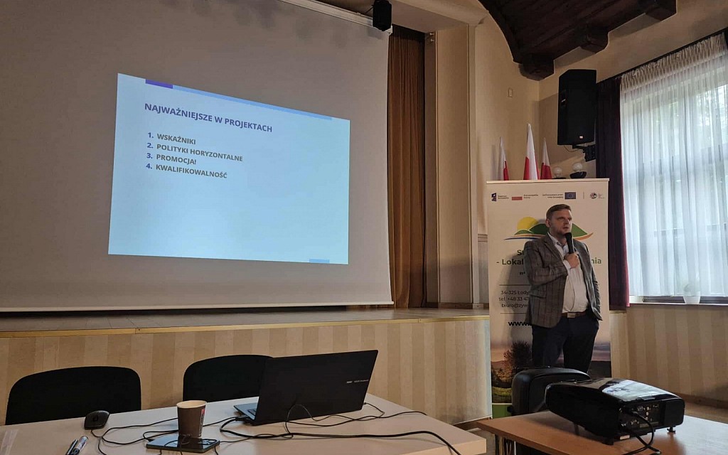 Prelegent stoi z mikrofonem obok ekranu, na którym wyświetlony jest slajd zatytułowany „Najważniejsze w projektach” z wypunktowanymi czterema zagadnieniami. Obok niego stoi roll-up LGD „Żywiecki Raj”.