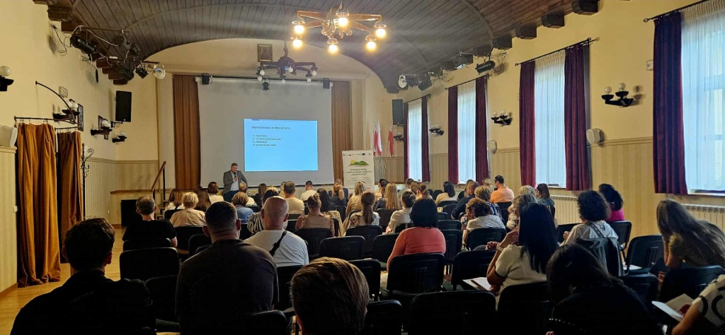Widok ogólny sali konferencyjnej z uczestnikami szkolenia siedzącymi w rzędach i słuchającymi prelekcji. W tle widoczny baner LGD oraz okna z zasłonami.