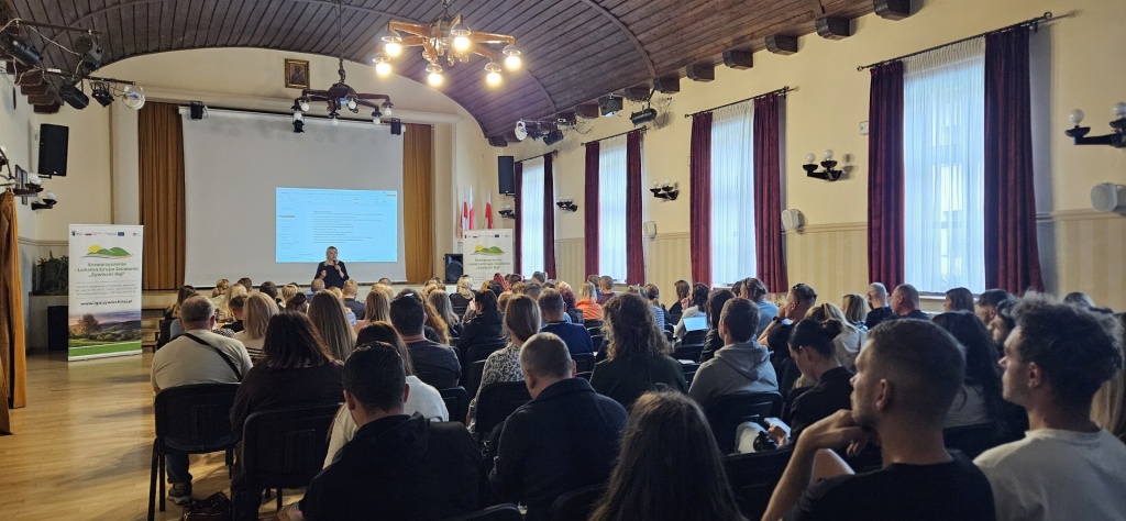Widok sali Gminnego Ośrodka Kultury w Łodygowicach podczas szkolenia LGD Żywiecki Raj. Prowadząca występuje przed liczną grupą uczestników, na ekranie wyświetlana jest prezentacja, a po bokach ustawione są roll-upy informacyjne.