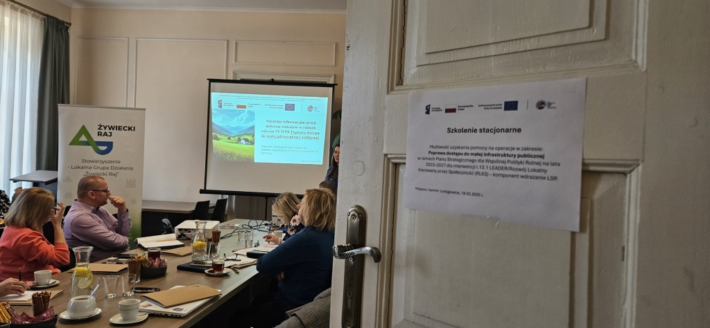 Foto 1 Zdjęcie przedstawia salę szkoleniową podczas spotkania informacyjnego organizowanego przez Stowarzyszenie Lokalna Grupa Działania „Żywiecki Raj”. Uczestnicy siedzą przy stole, słuchając prezentacji wyświetlanej na ekranie. Na pierwszym planie widoczna jest część drzwi z kartką informującą o temacie szkolenia – „Poprawa dostępu do małej infrastruktury publicznej”. W tle znajduje się roll-up z logo Żywiecki Raj.
