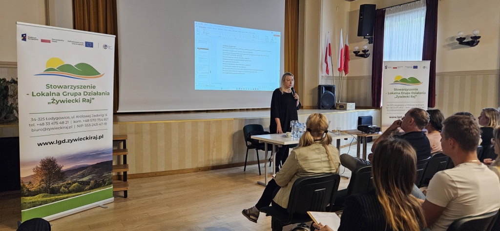 Prowadząca szkolenie przemawia do uczestników zgromadzonych w sali Gminnego Ośrodka Kultury w Łodygowicach. Obok widoczny jest ekran z prezentacją i roll-up LGD Żywiecki Raj.